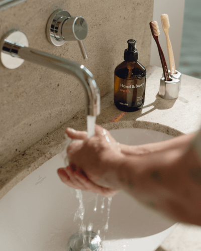 Person washing hands under running water in a sink with a bottle of arvessa.live's Hand & Body Soap bottle and toothbrushes in the background.